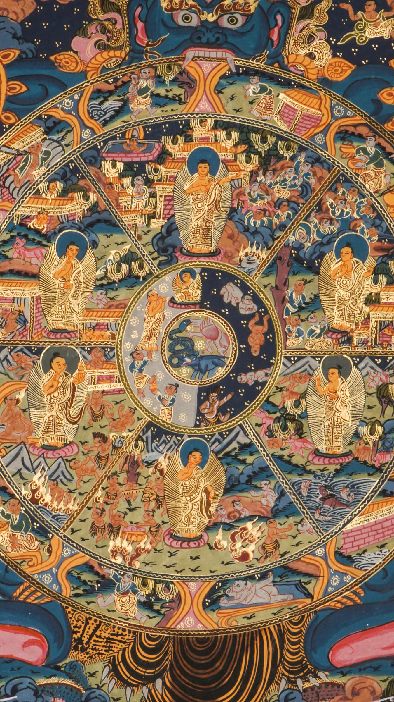 Detailed thangka-style painting showing a large circular wheel divided into multiple sections filled with small human figures, animals, buildings, and landscape scenes, surrounded by concentric decorative rings and held by a large blue multi-eyed figure with extended arms, set against a dark background with clouds, flames, and ornate gold, red, and blue patterns.