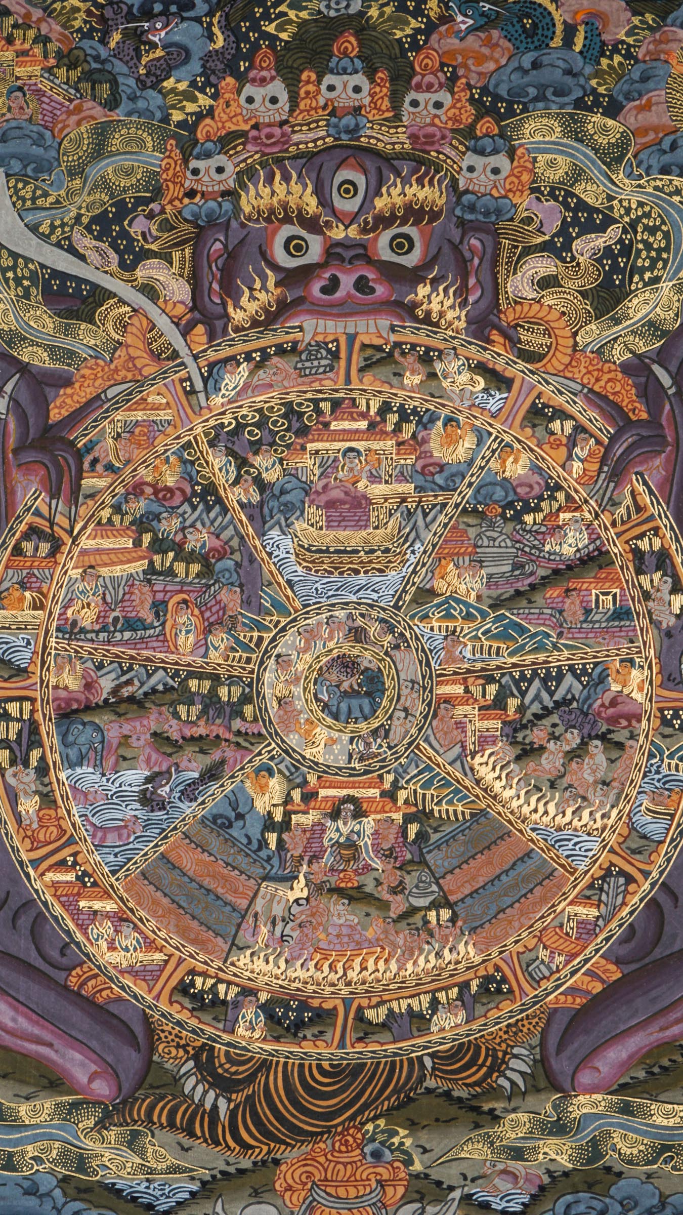 Detailed thangka-style painting showing a large circular wheel divided into multiple sections filled with small human figures, animals, buildings, and landscape scenes, surrounded by concentric decorative rings and held by a large blue multi-eyed figure with extended arms, set against a dark background with clouds, flames, and ornate gold, red, and blue patterns.