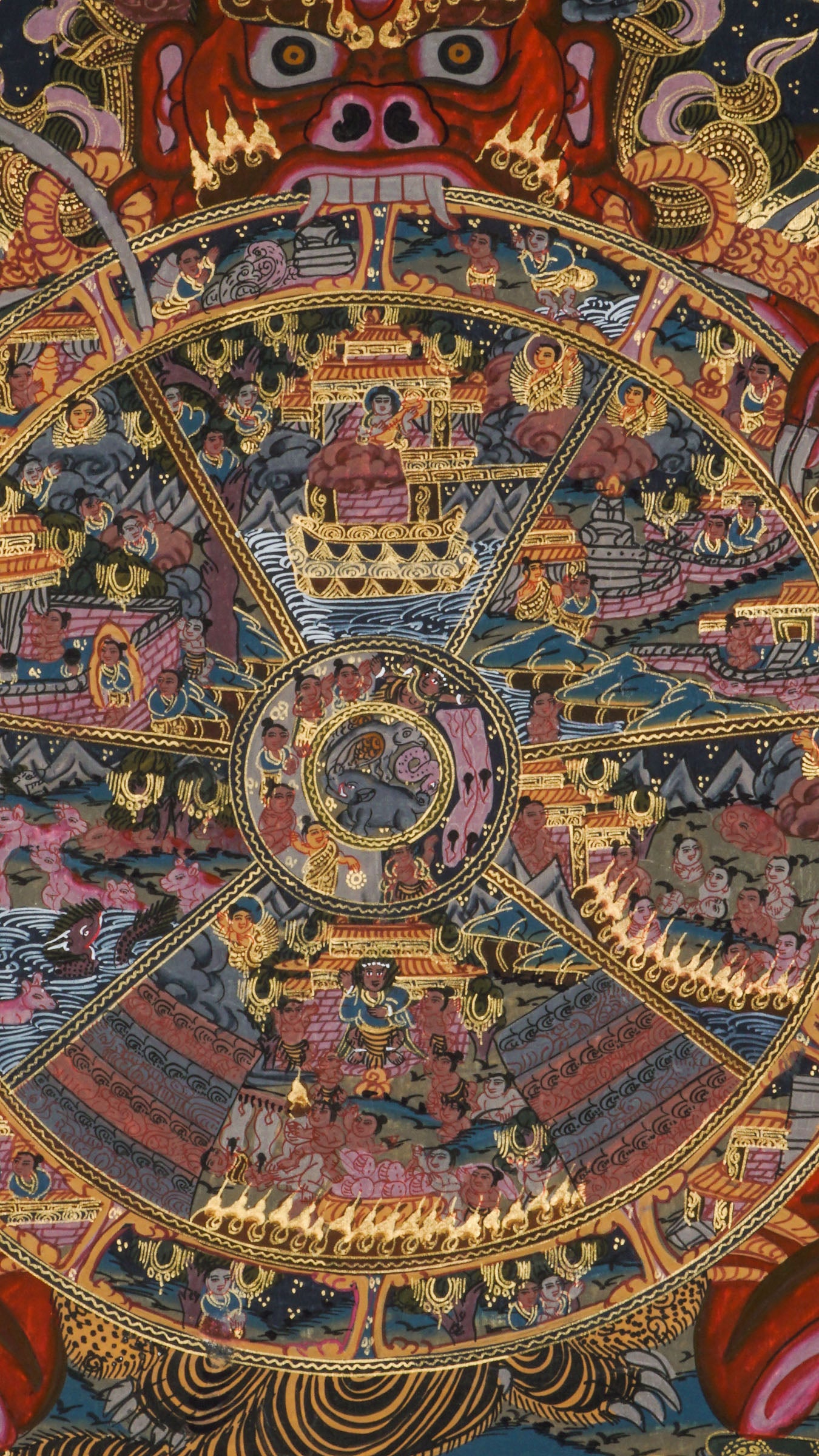 Detailed thangka-style painting showing a large circular wheel divided into multiple sections filled with small human figures, animals, buildings, and landscape scenes, surrounded by concentric decorative rings and held by a large blue multi-eyed figure with extended arms, set against a dark background with clouds, flames, and ornate gold, red, and blue patterns.