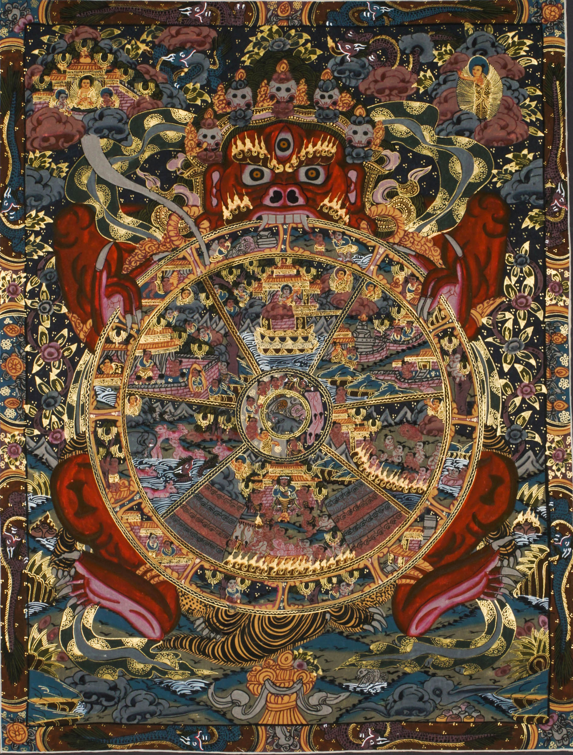 Detailed thangka-style painting showing a large circular wheel divided into multiple sections filled with small human figures, animals, buildings, and landscape scenes, surrounded by concentric decorative rings and held by a large blue multi-eyed figure with extended arms, set against a dark background with clouds, flames, and ornate gold, red, and blue patterns.