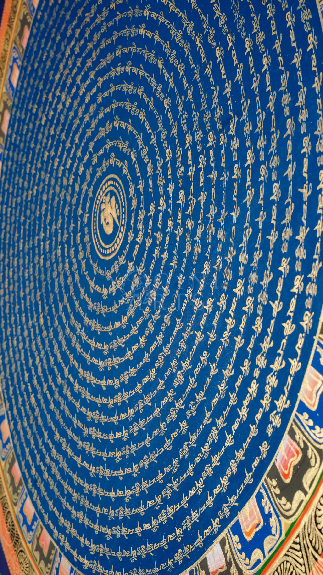 Blue Om Mantra Mandala thangka featuring sacred Om symbol surrounded by concentric circles of gold Tibetan mantra calligraphy and floral corner motifs.