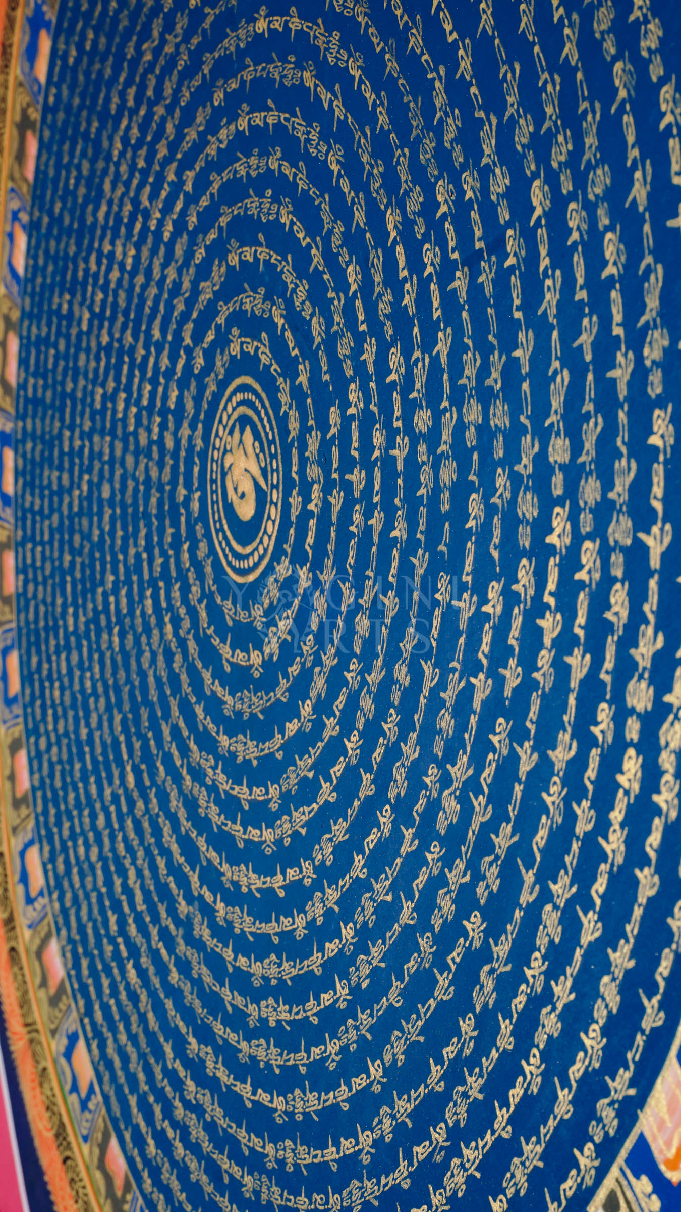 Blue Om Mantra Mandala thangka featuring sacred Om symbol surrounded by concentric circles of gold Tibetan mantra calligraphy and floral corner motifs.