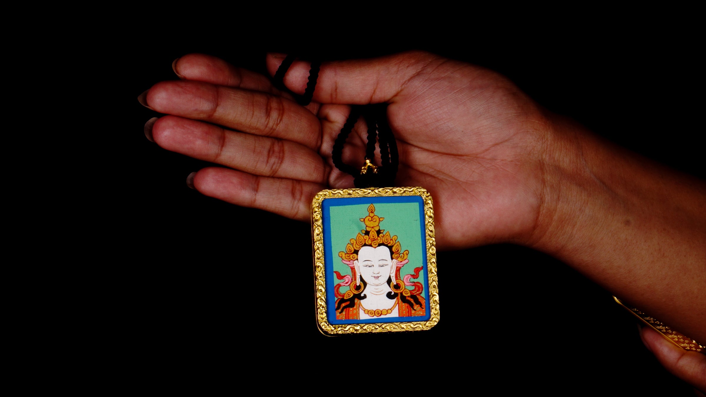 Square gold-framed Vajrasattva pendant with blue background, held in an open hand against a black backdrop.