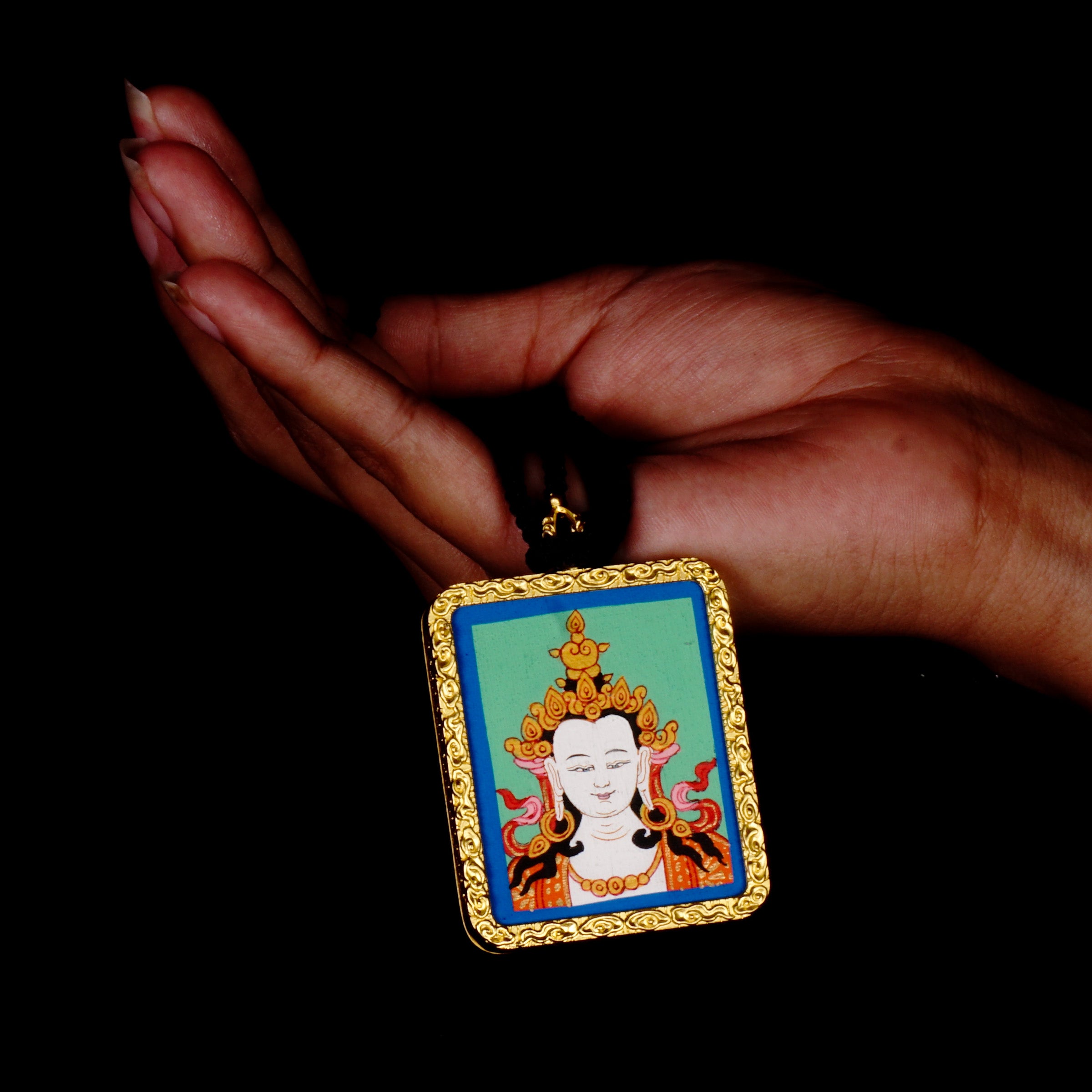 Square gold-framed Vajrasattva pendant with blue background, held in an open hand against a black backdrop.