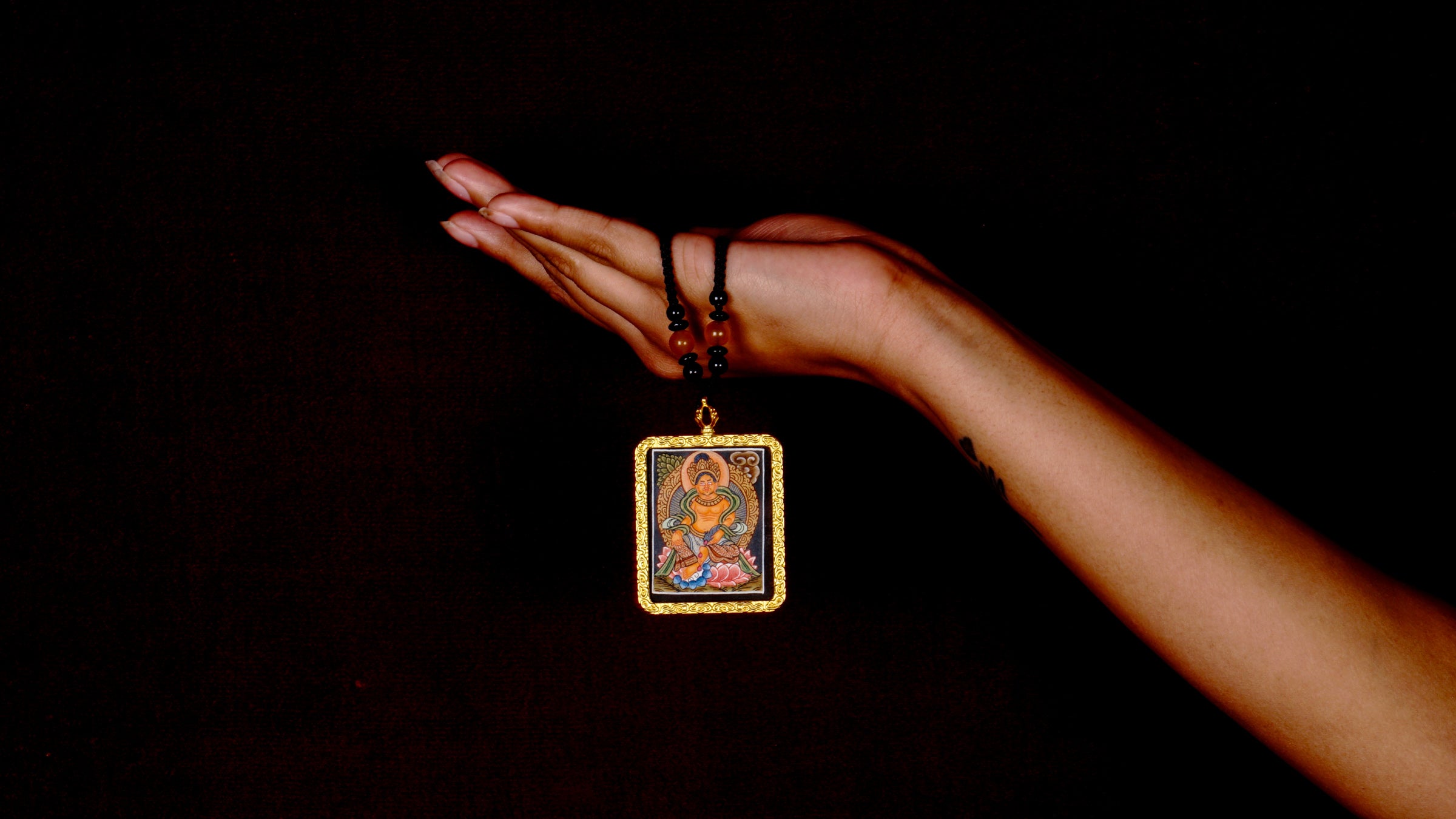 Hand-painted Zambala miniature Thangka pendant in gold-accented copper Ghau locket displayed hanging from black beaded cord.