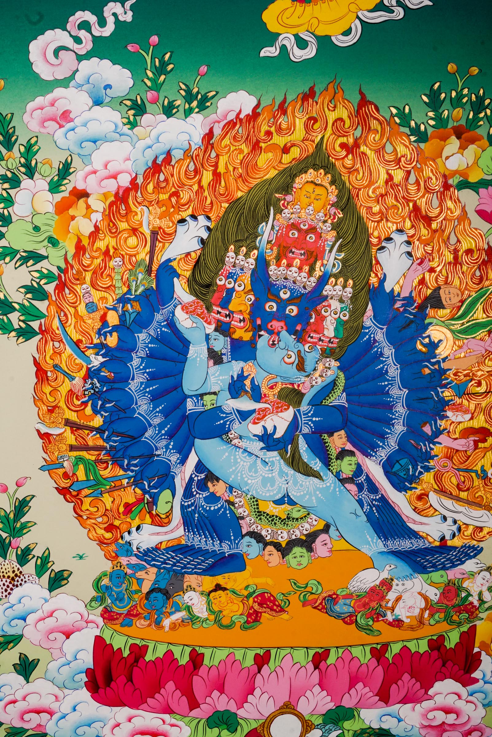 Traditional Tibetan thangka painting depicting Yamantaka Shakti in dynamic embrace, surrounded by flames, clouds, and abundant floral motifs, rendered in vivid colors against a green and turquoise landscape.