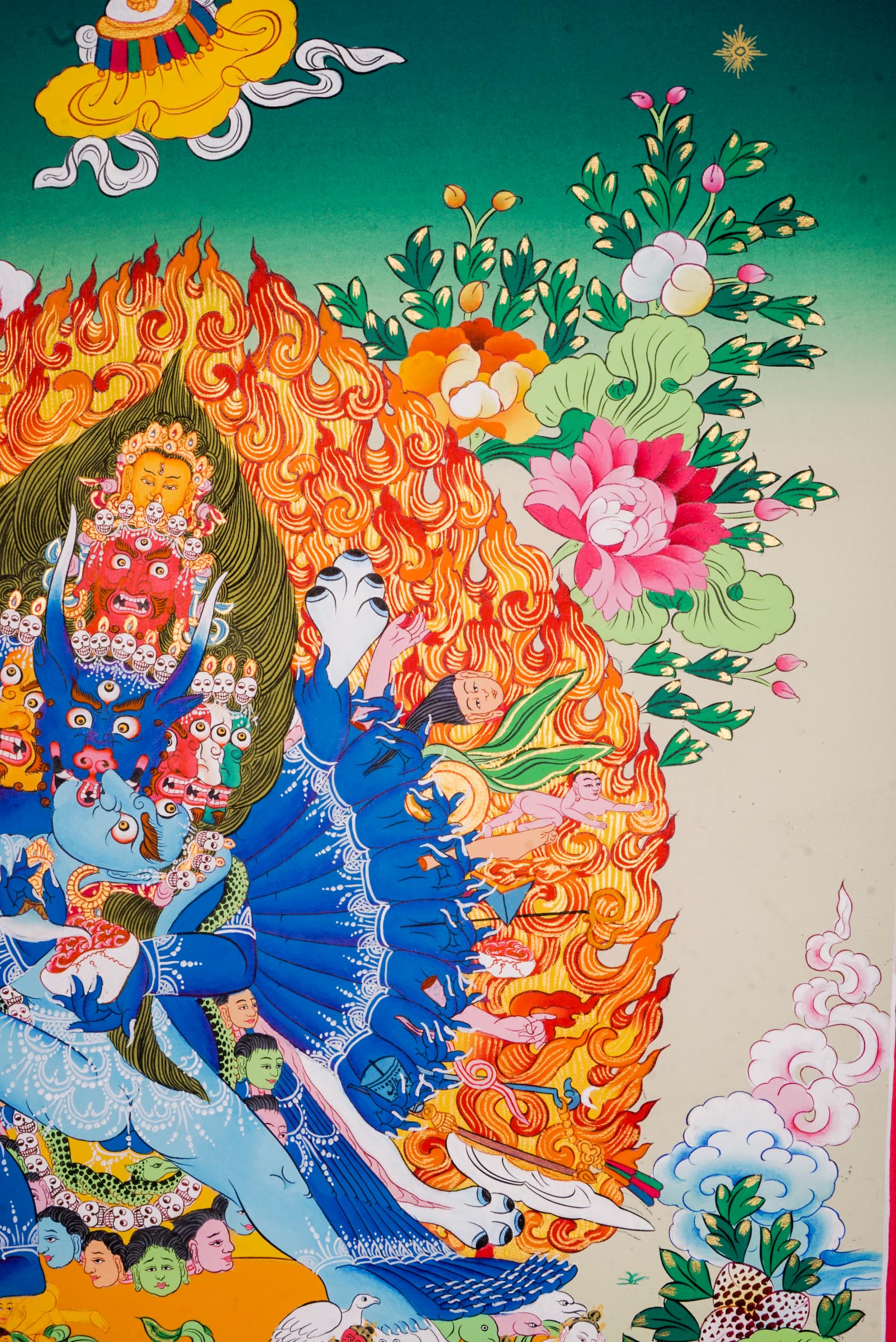 Traditional Tibetan thangka painting depicting Yamantaka Shakti in dynamic embrace, surrounded by flames, clouds, and abundant floral motifs, rendered in vivid colors against a green and turquoise landscape.