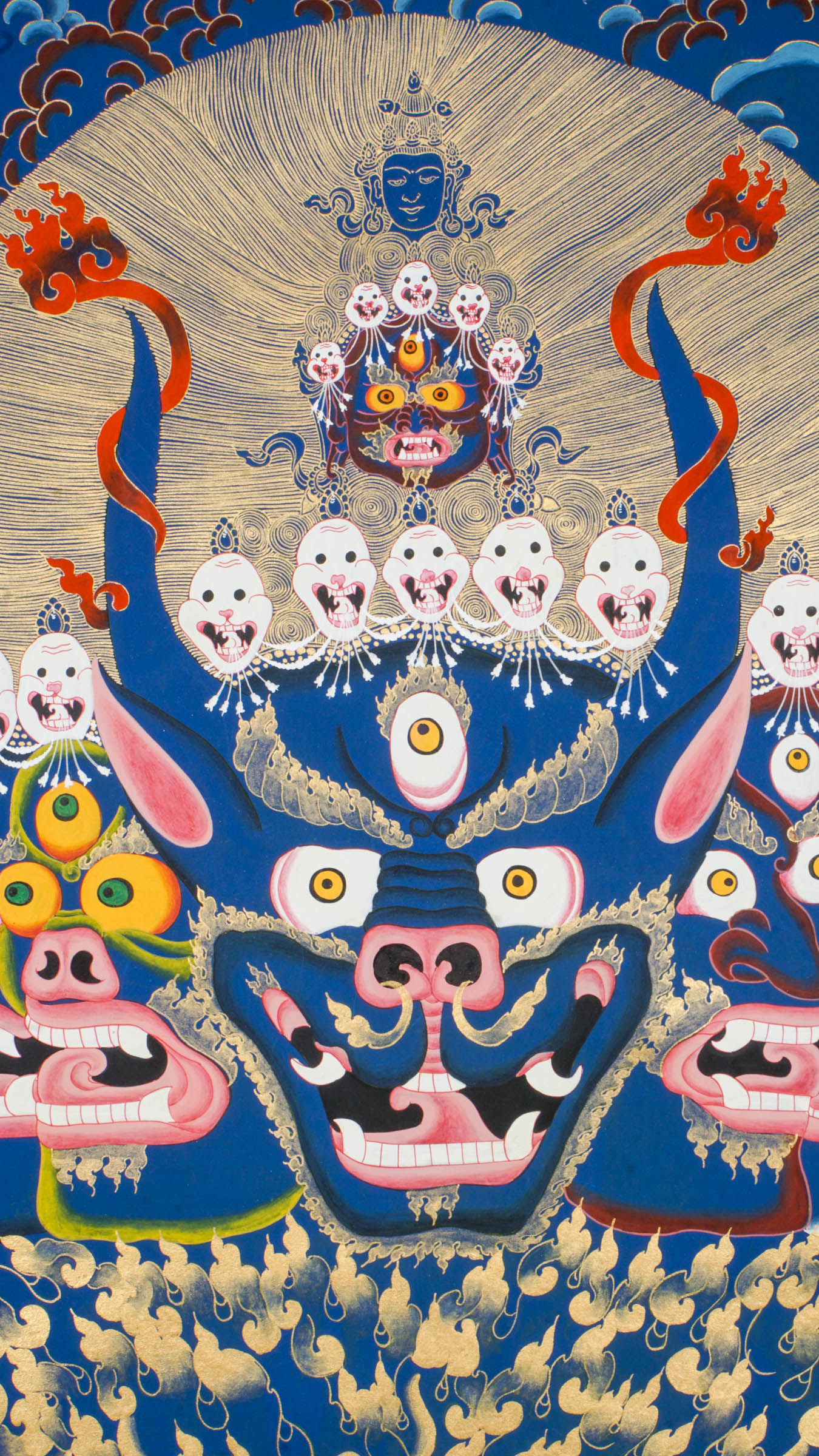 Thangka painting showing a large blue multi-faced figure with numerous eyes and open mouths, crowned by smaller faces and skull motifs, set against a gold halo and stylized cloud background.