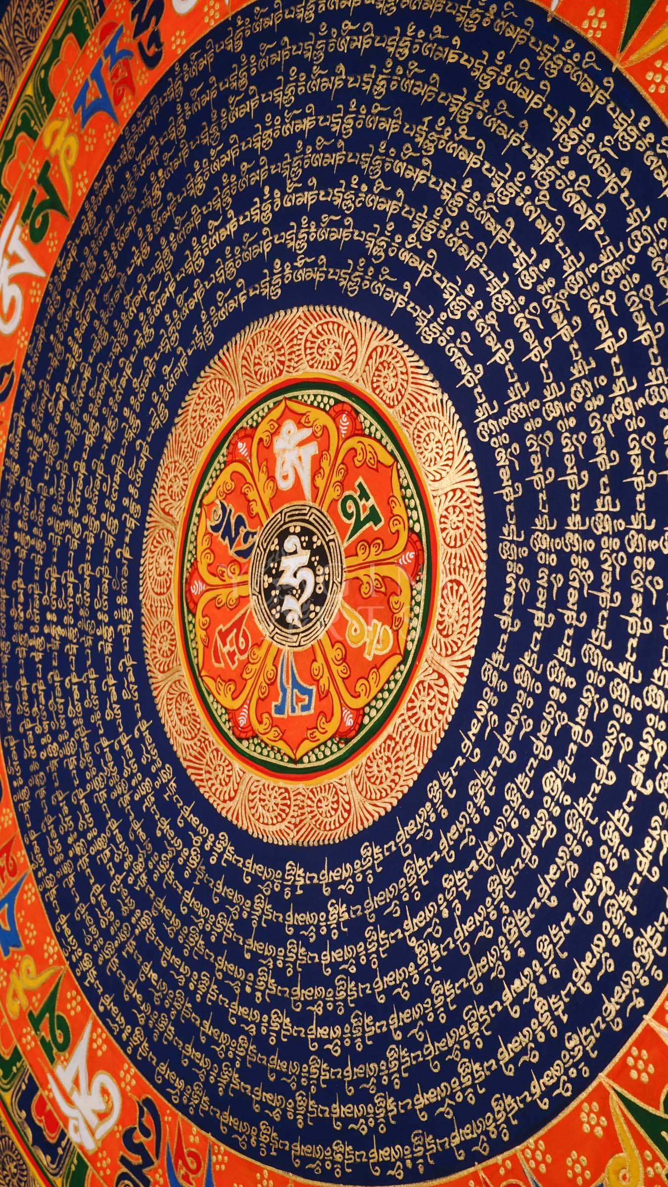 Tibetan mantra mandala thangka with blue background, gold script rings, colorful outer mantra band, and floral corner decorations.