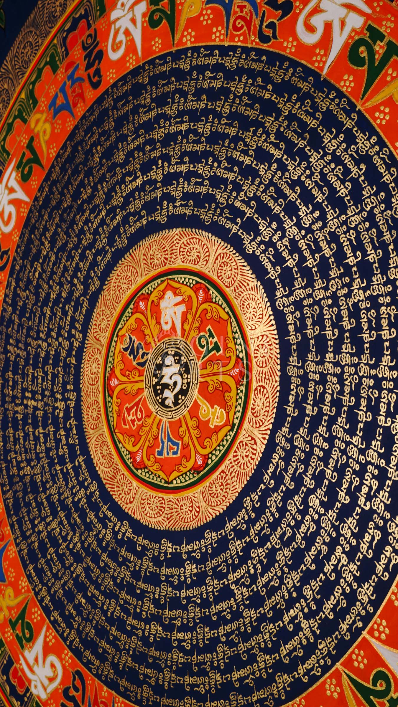 Tibetan mantra mandala thangka with blue background, gold script rings, colorful outer mantra band, and floral corner decorations.