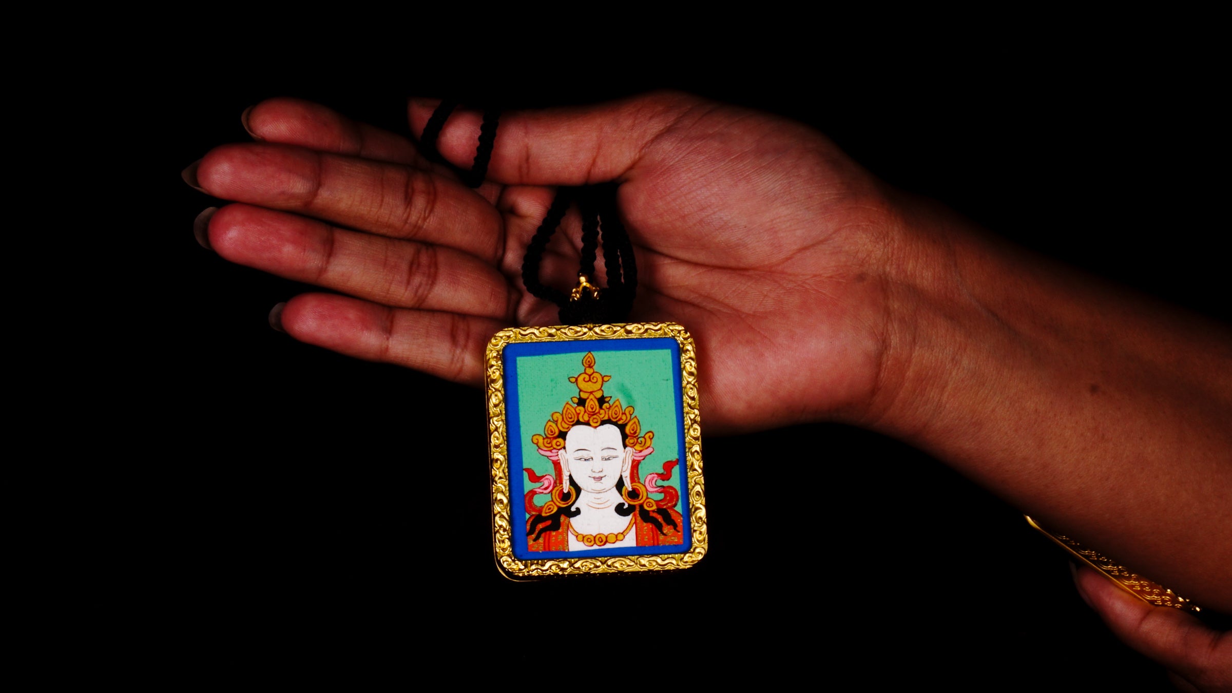 Square gold-framed Vajrasattva pendant with blue background, held in an open hand against a black backdrop.
