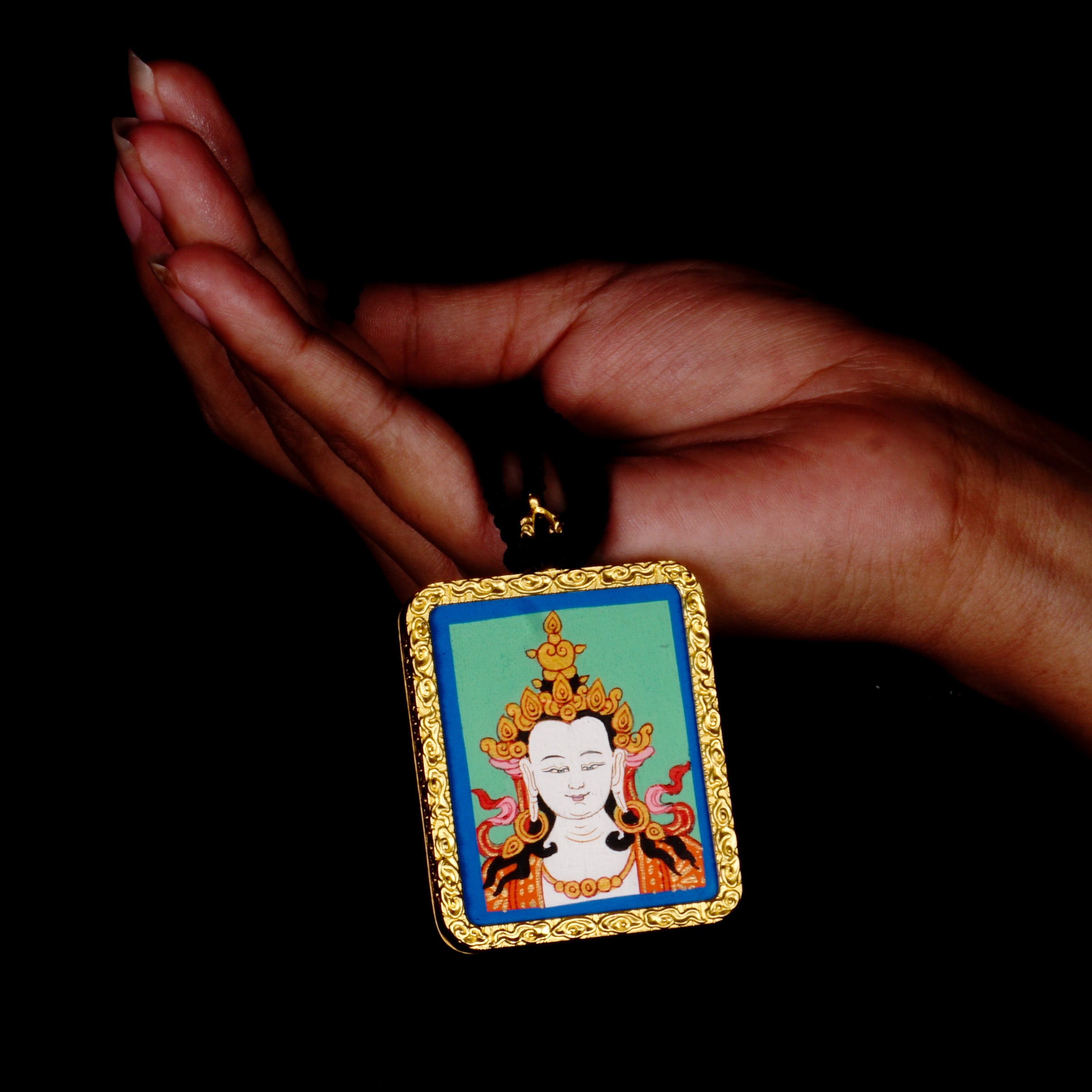Square gold-framed Vajrasattva pendant with blue background, held in an open hand against a black backdrop.
