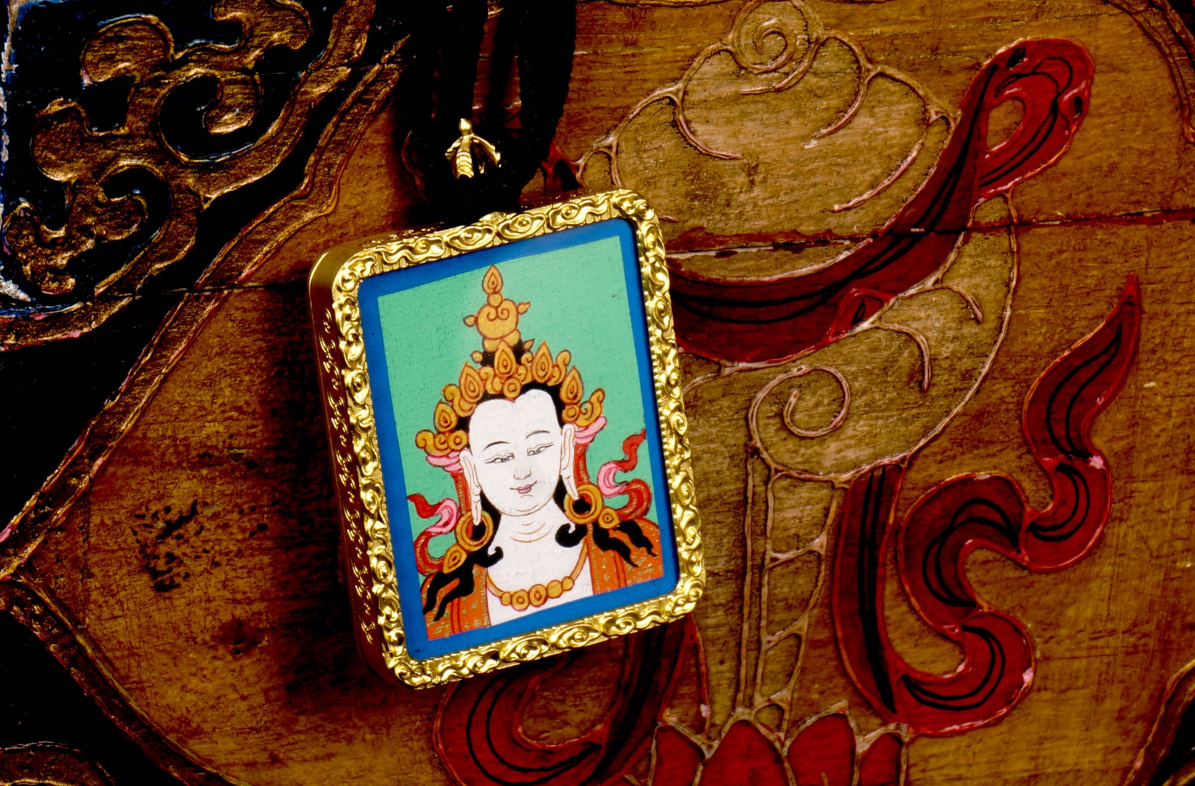 Square gold-framed Vajrasattva pendant with blue background, held in an open hand against a black backdrop.
