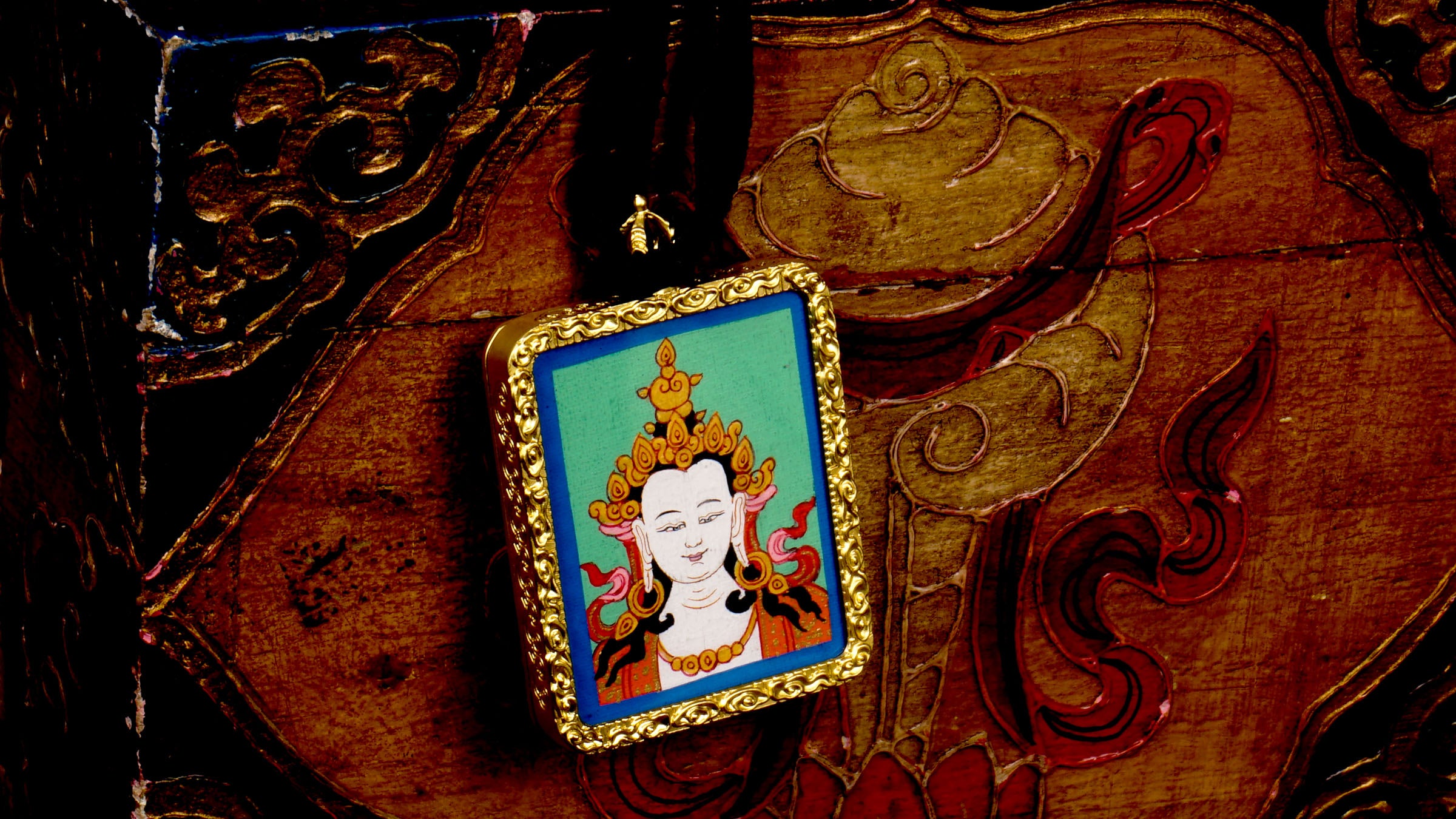 Square gold-framed Vajrasattva pendant with blue background, held in an open hand against a black backdrop.
