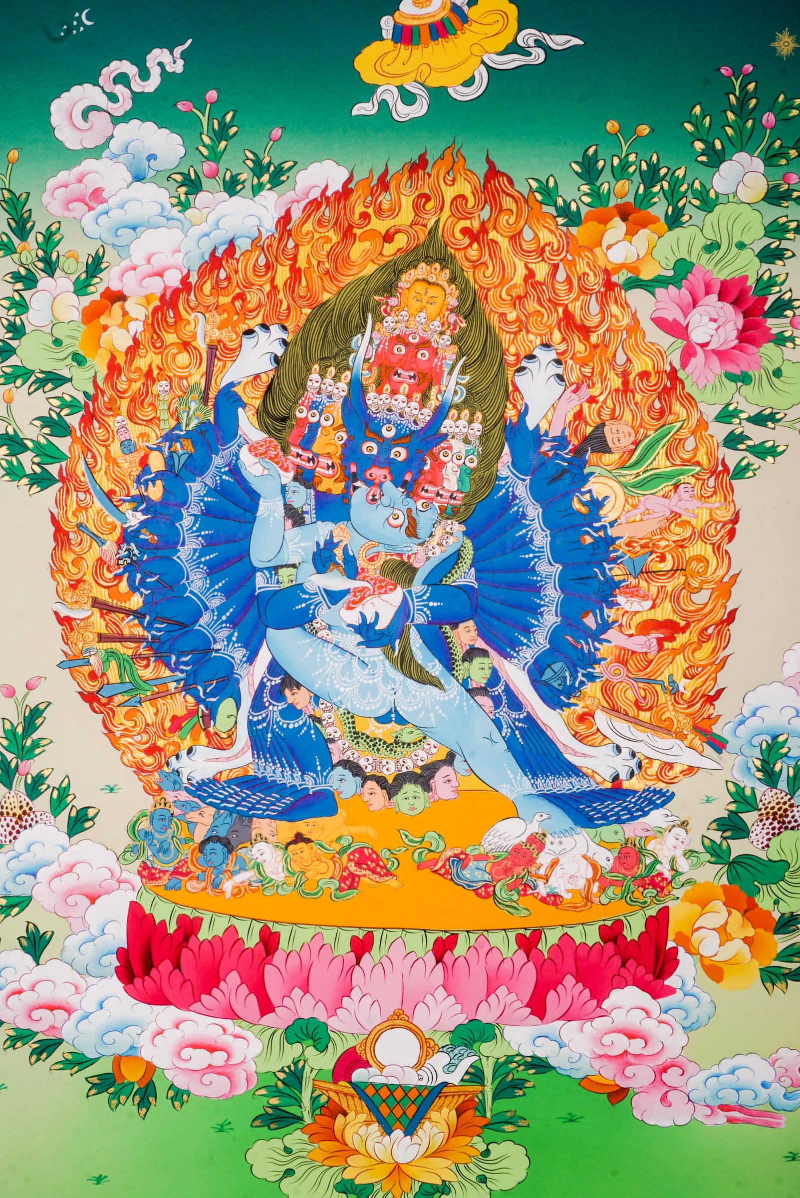 Traditional Tibetan thangka painting depicting Yamantaka Shakti in dynamic embrace, surrounded by flames, clouds, and abundant floral motifs, rendered in vivid colors against a green and turquoise landscape.