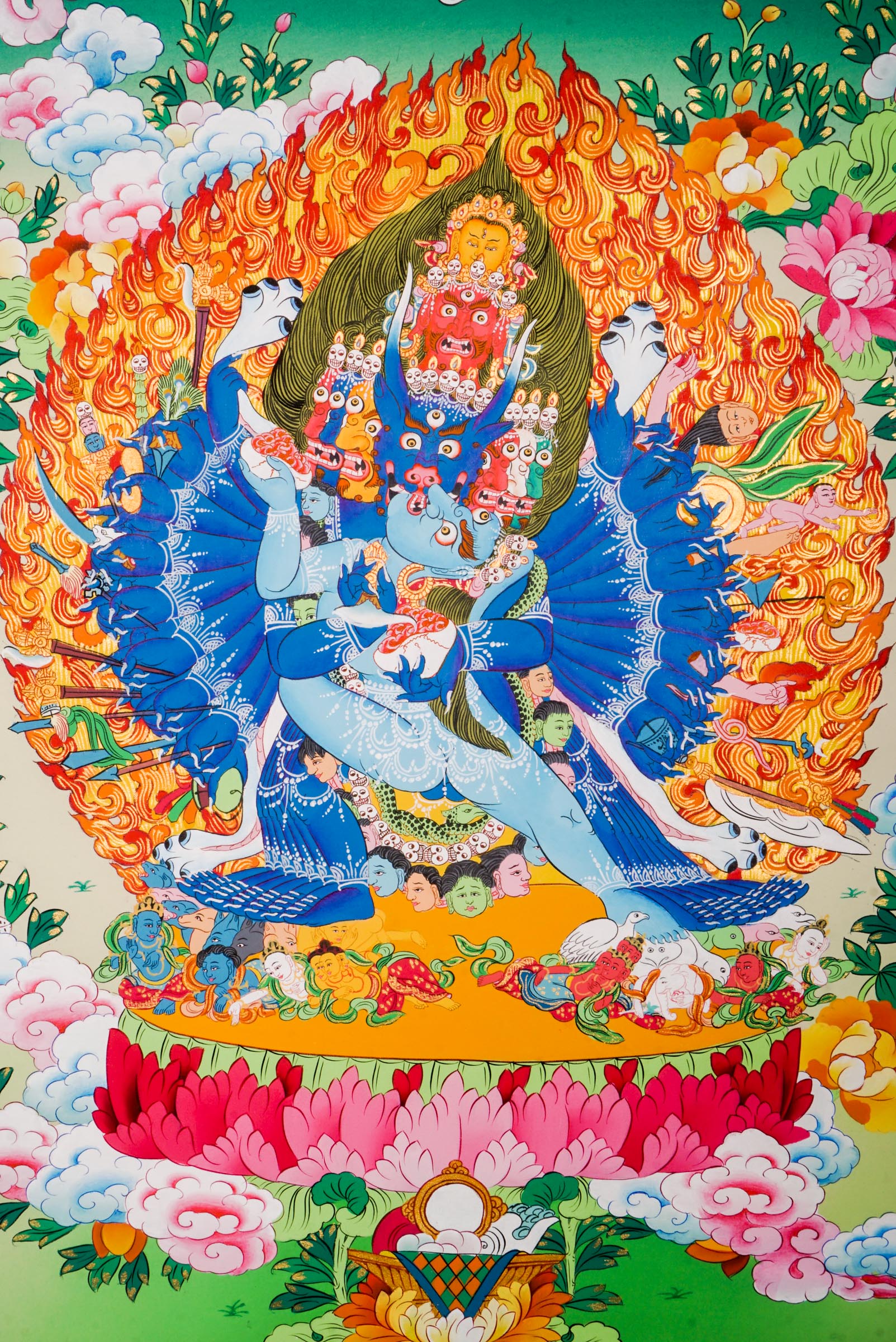 Traditional Tibetan thangka painting depicting Yamantaka Shakti in dynamic embrace, surrounded by flames, clouds, and abundant floral motifs, rendered in vivid colors against a green and turquoise landscape.