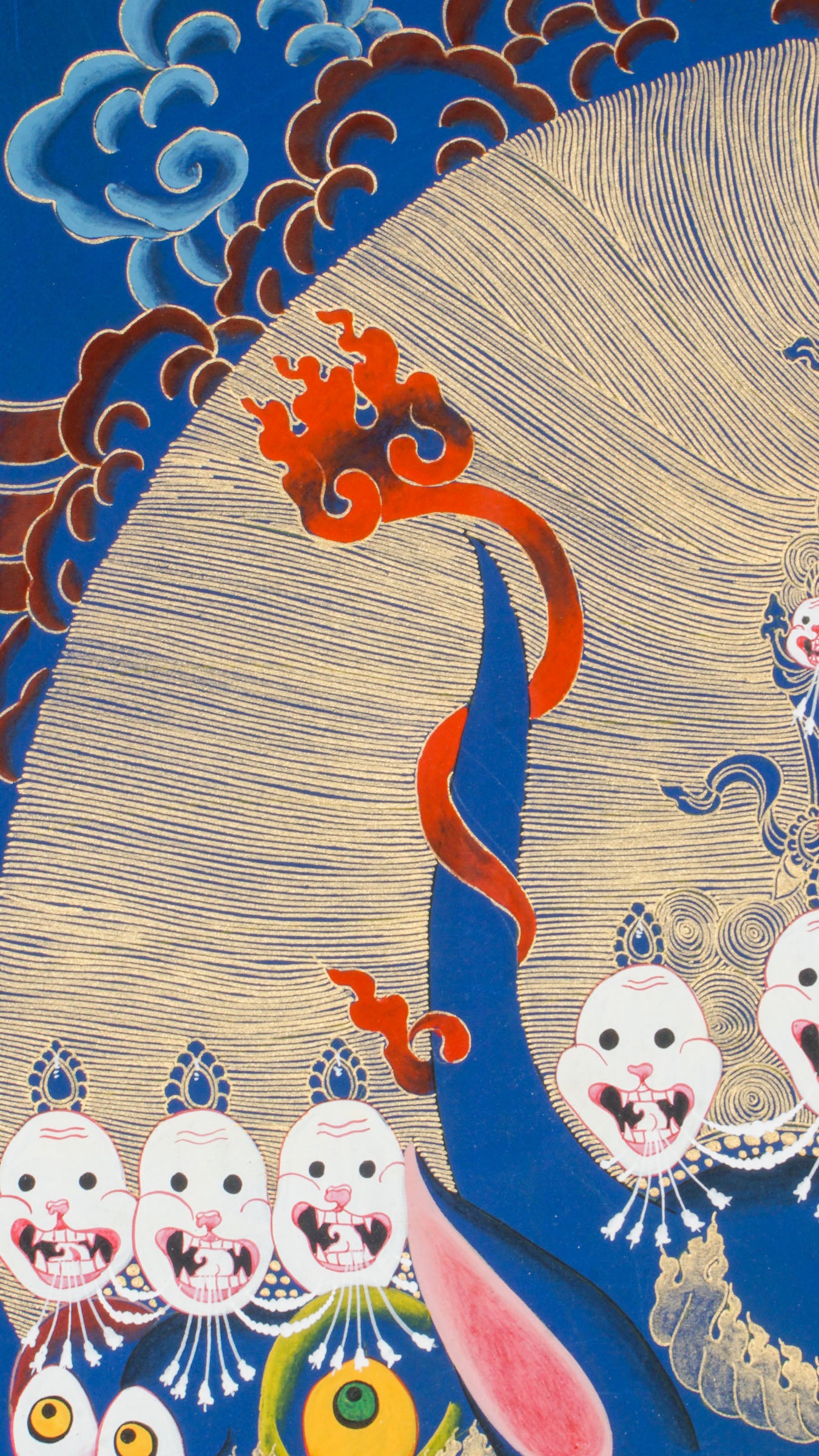 Thangka painting showing a large blue multi-faced figure with numerous eyes and open mouths, crowned by smaller faces and skull motifs, set against a gold halo and stylized cloud background.