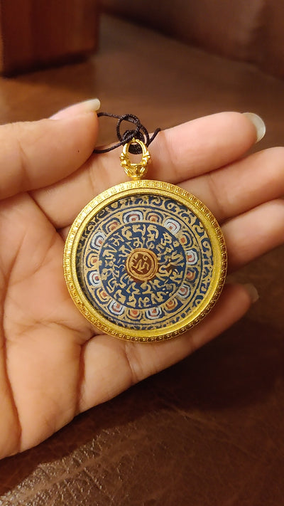 Hand-painted Mantra Mandala Ghau pendant featuring the sacred OM symbol and Tibetan Buddhist mantras in a handcrafted gold-tone frame.