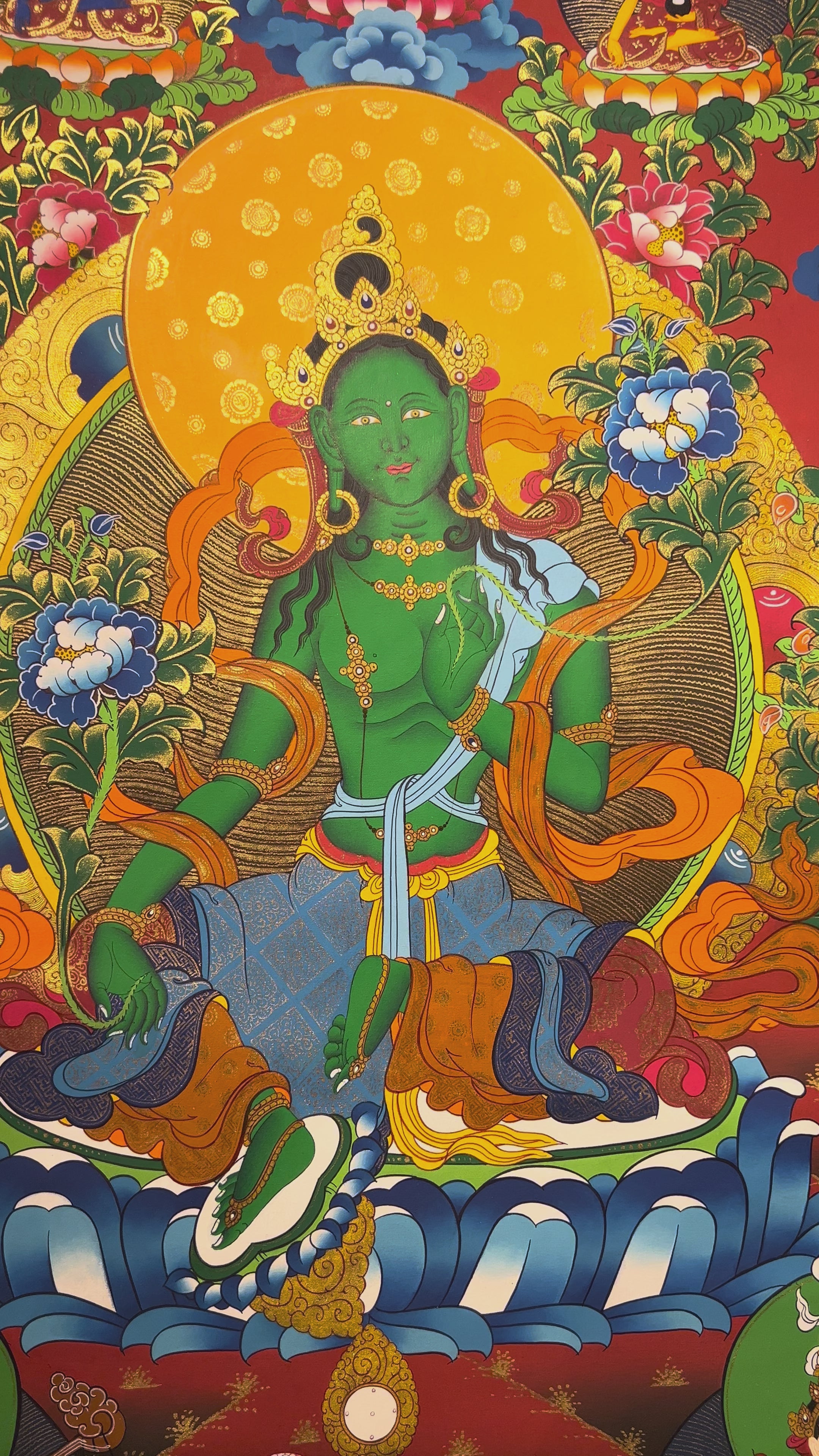 Green Tara thangka featuring the bodhisattva seated on a lotus throne with lotus flowers, surrounded by celestial figures and vibrant Himalayan landscape.