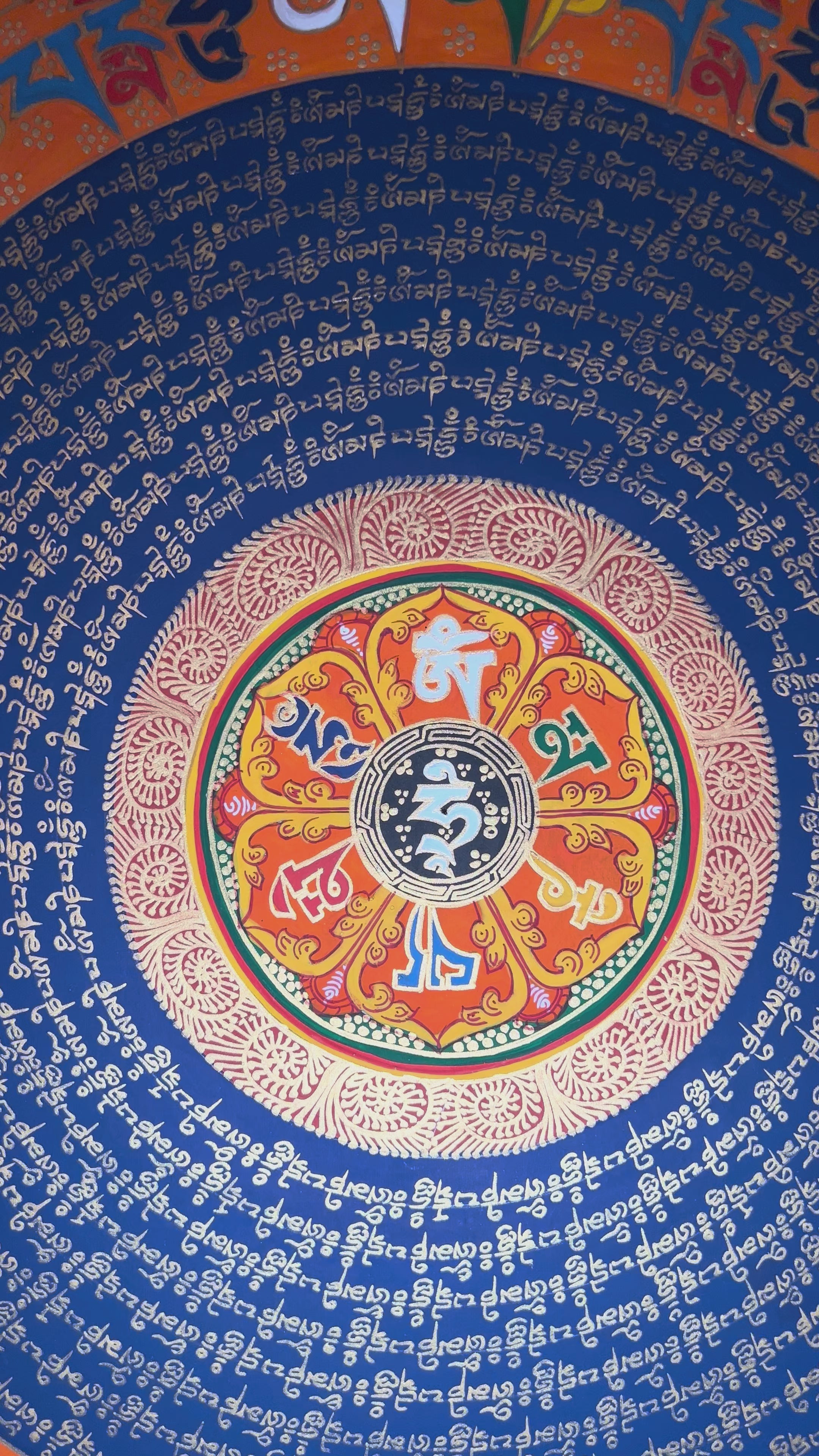 Tibetan mantra mandala thangka with blue background, gold script rings, colorful outer mantra band, and floral corner decorations.